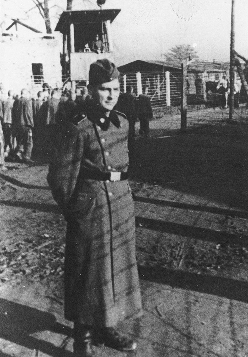 Guard outside of Auschwitz entrance. Prisoners marching in the back.