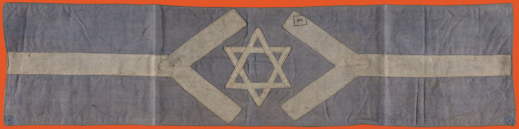 Armband with a light blue background and a horizontal white line crossing it. At the center the horizontal line opens up in two diagonal lines and a white star of david is at the center