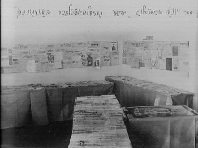 Black and white photo of a room with tables covered in tablecloth, with materials on top. The walls have materials taped on them - photos and other printed materials.