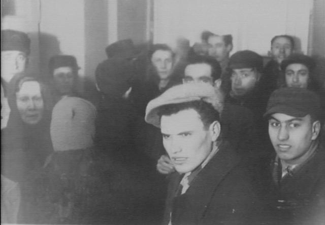 Black and white photo of a crowded room. People are depicted from the chest up. A man is at the center. He is white and wears a hat.