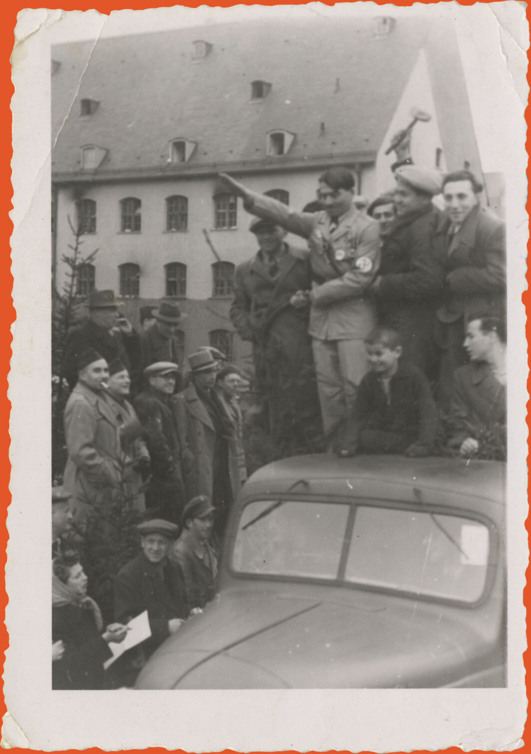 Black and white photo. A building with 2 or 3 floors is in the background. The street is crowded and on the front, to the right, is a car with people on top of it. Right in the middle is a man dressed up as Hitler making the arm salute.