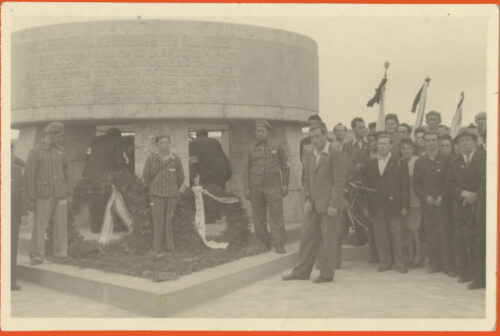 Black and white photo of 3 people in front of a circular structure. They wear striped uniforms. On the left there is a crowd.