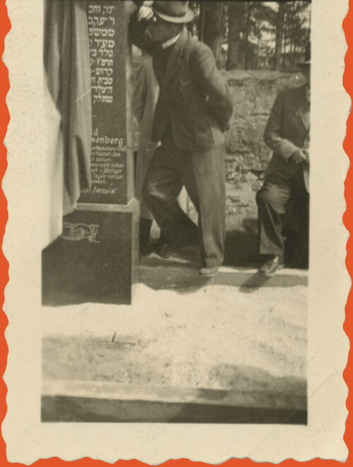 Black and white photo of a man leaning on a monument with a falg over half of it. His arm lays on the monument and his head is resting over his arm.