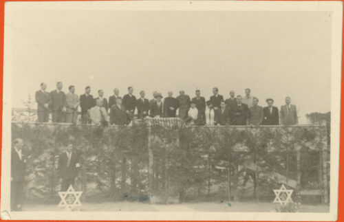 Black and white photo of a group of people orderly organized on the top of what looks like a wooden structure. Two people stand below on the sides, each with a star of david in the ground standing in front of them.