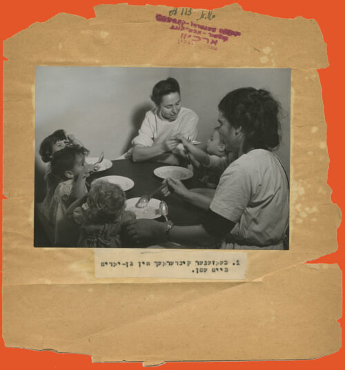 Black and white photo glued to a paper with torn edges and a caption typed in Yiddish below. The photo shows people sitting at a table and eating. Two women to the right side and three children to the left.