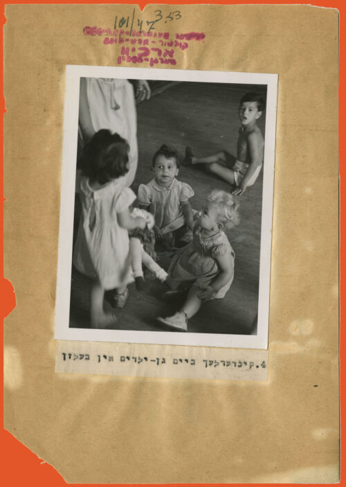 Black and white photo glued to a paper with torn edges and a typed caption in Yiddish below. The picture displays a little girl with her back to the camera to the right of the picture, next to a person who we can only see is wearing a skirt as they are shown only from the waist down. Two children are sitting on the floor right in front of them facing the camera, and another is at the back, also sitting on the floor and facing the camera.