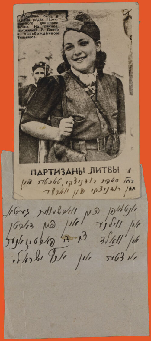 Black and white portrait of a woman from the waist up. She wears a military uniform, with hat, is smiling and looking to the side, and has a bag over her shoulder while she holds the strap. Below is a piece of paper with text in Yiddish.