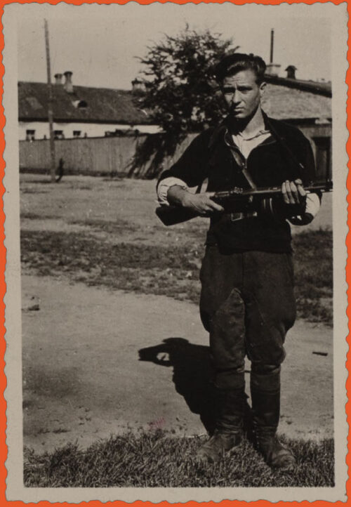 Black and white photo. A man stands to the right of the picture, in front of what looks like a dirt road. We can see a wall on the back and the roof of a one-story building. He wears a dark half-zip sweater, with the zipper open, over a light colored shirt, belt on top of both, dark culottes, and high boots. He holds a rifle.