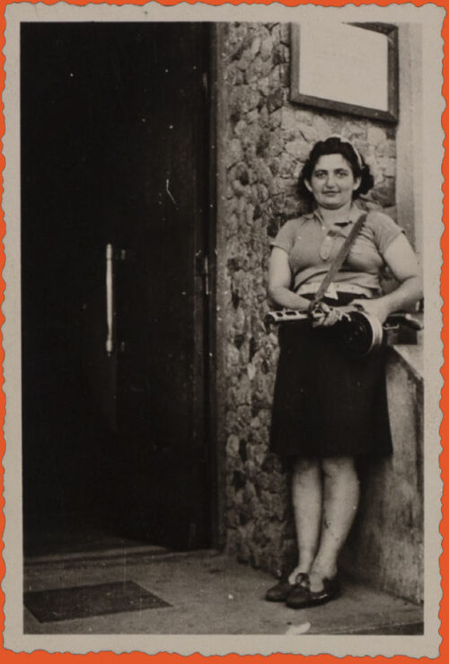 Black and white photo. A woman stands next to an open door, in front of a building, outside. She wears a short-sleeve shirt, knee-length dark skirt, and dark low-heel untied mary-janes. Her dark hair hits her shoulders and she is wearing a headband. She holds a big gun, possibly a rifle.