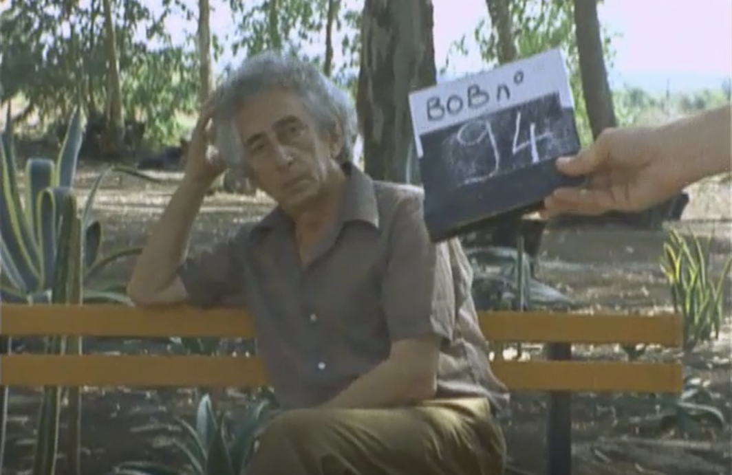 Photo of a man with gray hair wearing a brown short sleeve button up shirt and olive pants, sitting in a wooden bench outside, in front of trees with green leaves.